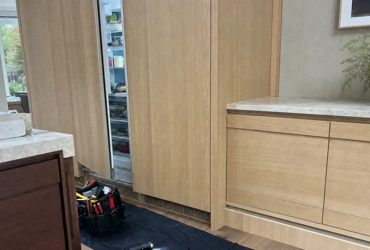 Pale oak fitted kitchen with a tall integrated cabinet standing open at the left and a cordless drill and tool bag on the floor